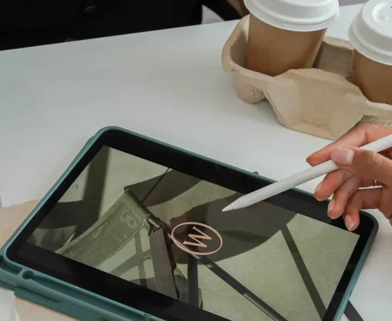 Hand using stylus on tablet displaying branded design, with takeaway coffee cups in background, symbolising Marketing Valley’s collaborative email marketing process.
